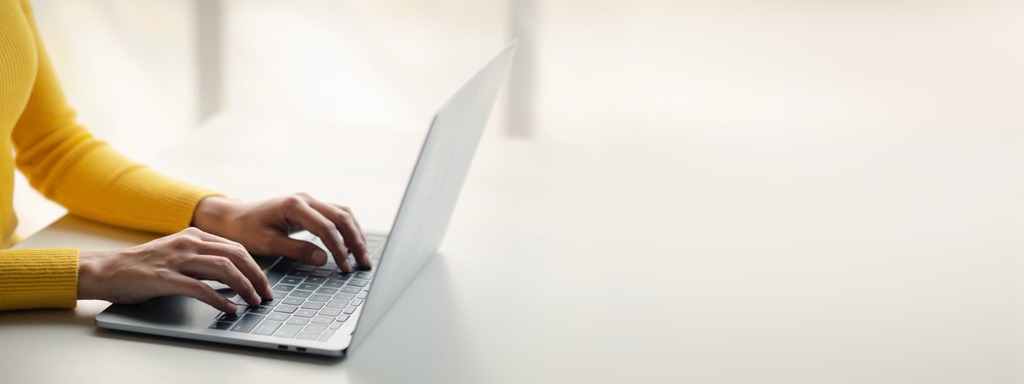 A person typing on a laptop sitting on a white table. Focus is on arms wearing a yellow sweater.