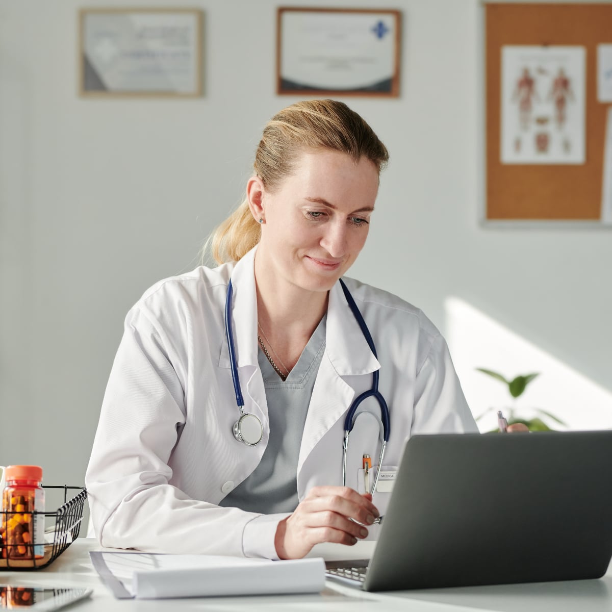 A woman doctor with blonde hair sitting at a desk using a laptop. She is wearing a lab coat and has a stethoscope. 