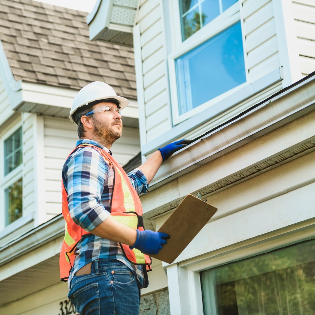 A home service worker wearing a hard hat and vest inspecting a home roof.