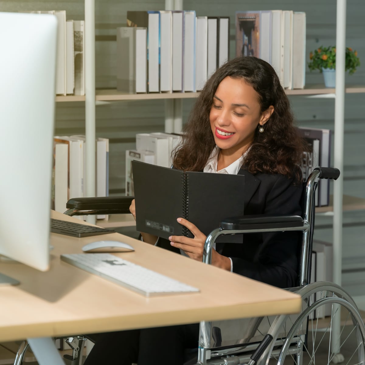 A young professional woman writing in a note pad. She is sitting in a wheel chair at a desk with a computer.