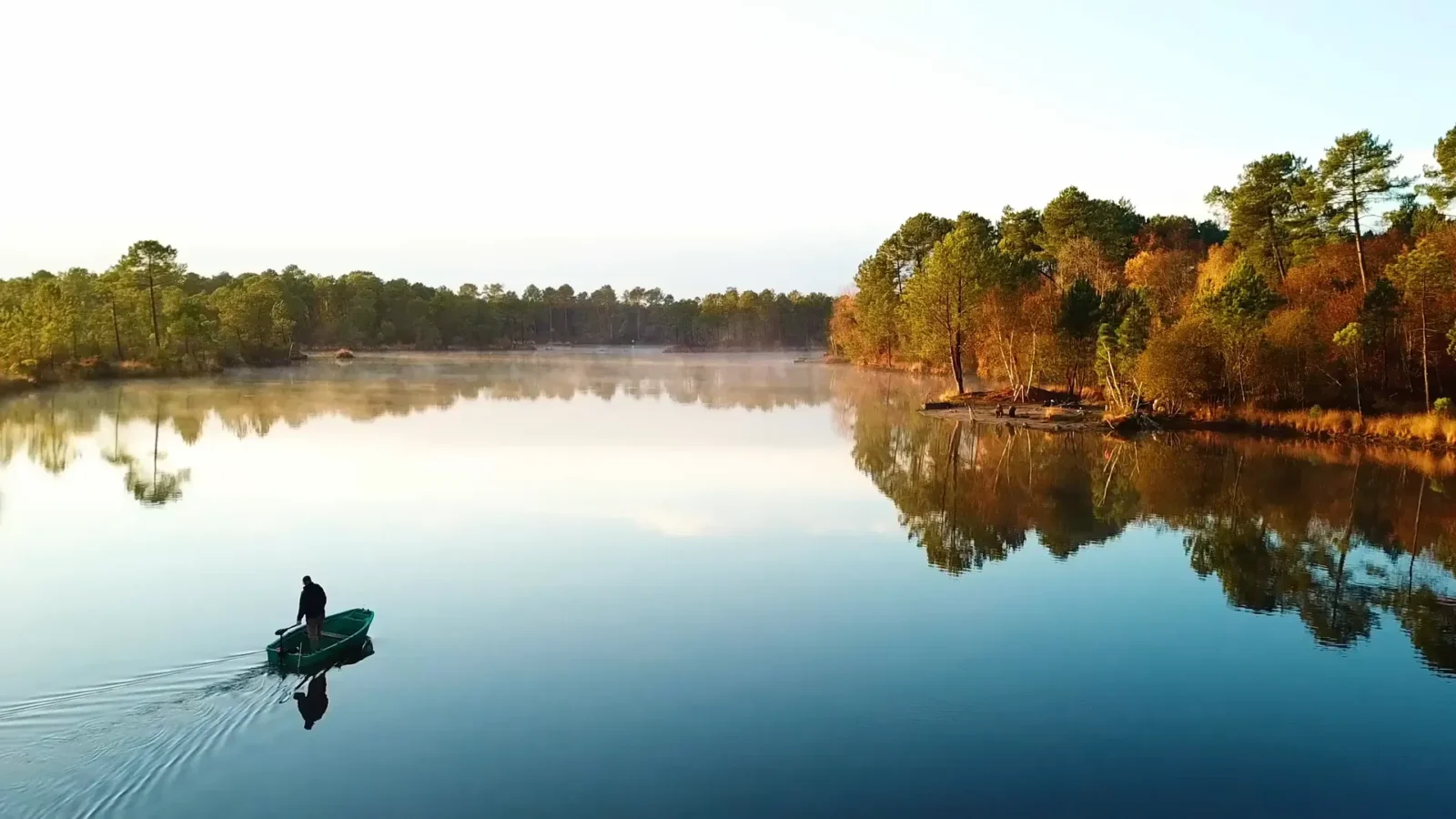 A person steers a small green boat across a calm lake, surrounded by a dense forest showing bright autumn colors reflecting in the water.