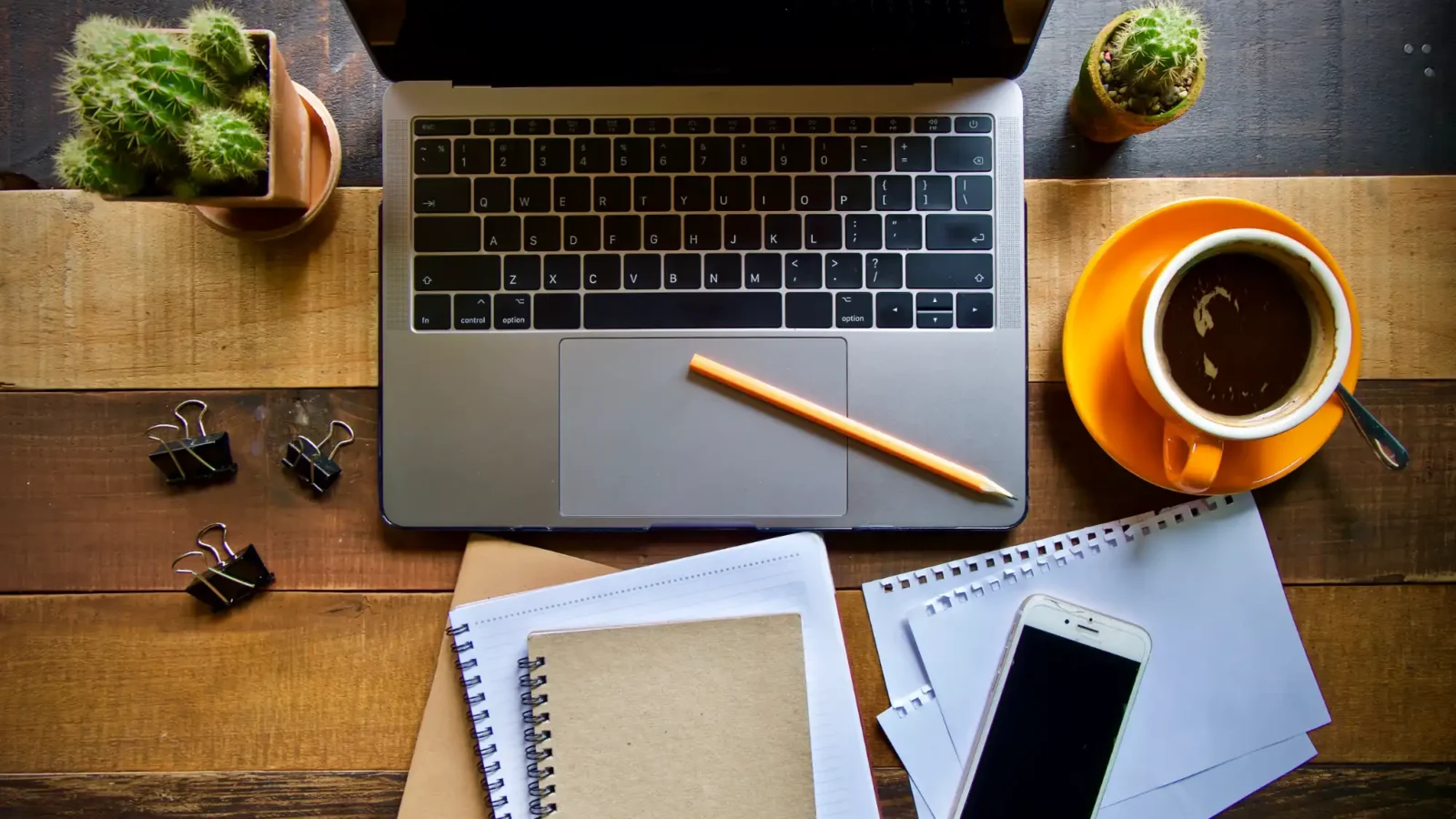A top view of a work desk featuring a laptop, notebooks, a smartphone, and an orange cup of coffee on a rustic wooden surface.