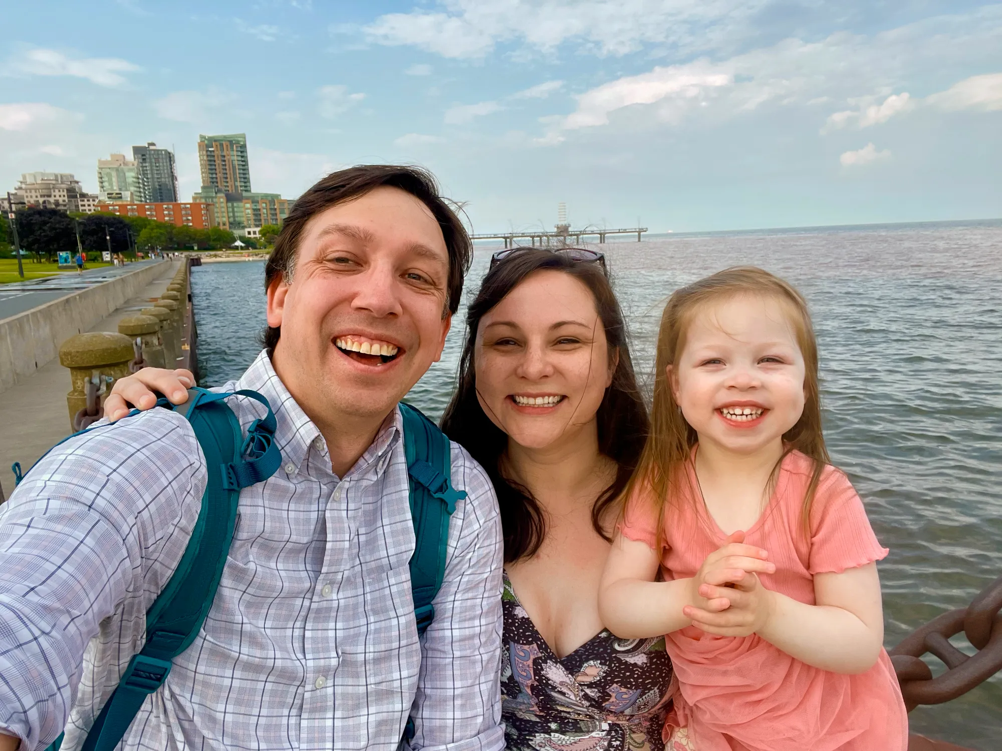 The Abalos family—a man with a backpack, a woman, and a young girl—are taking a joyful selfie on a pier overlooking a waterfront Toronto skyline.
