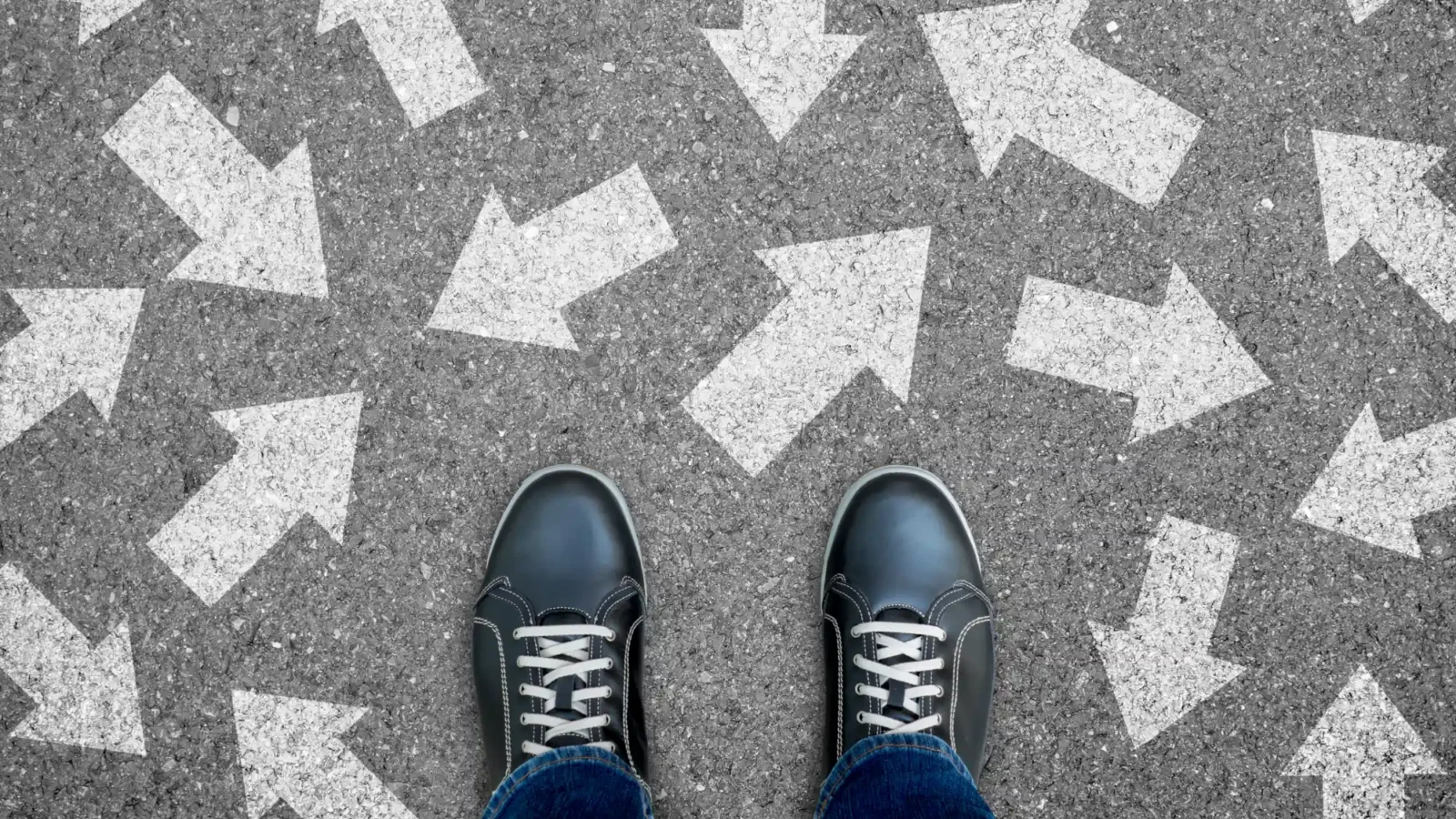 A person's black shoes stand on gray asphalt painted with large white arrows pointing in contradictory directions.