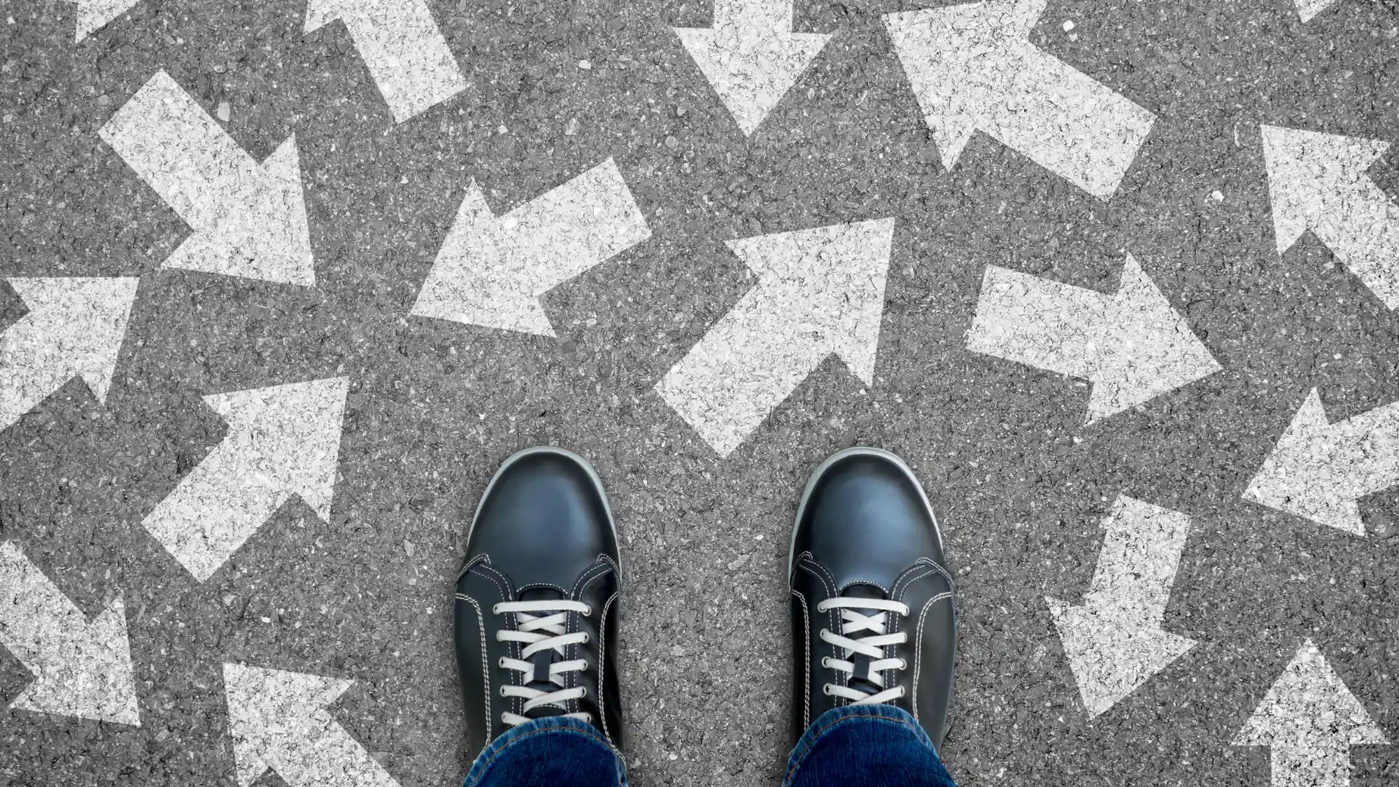 A person's black shoes stand on gray asphalt painted with large white arrows pointing in contradictory directions.
