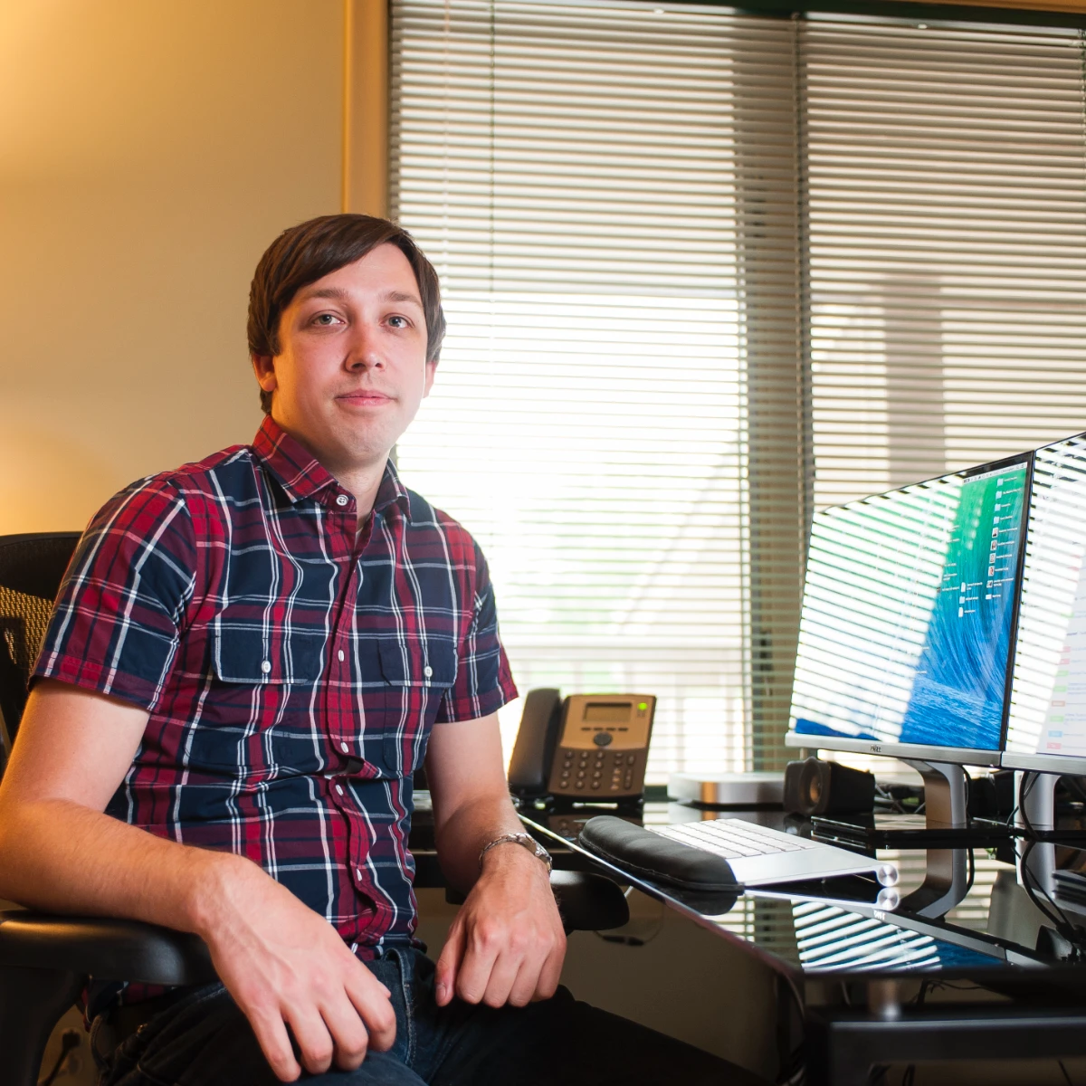 Chris, a professional, sits in an office with dual computer monitors and a multi-line phone. He wears a plaid shirt and looks forward calmly.