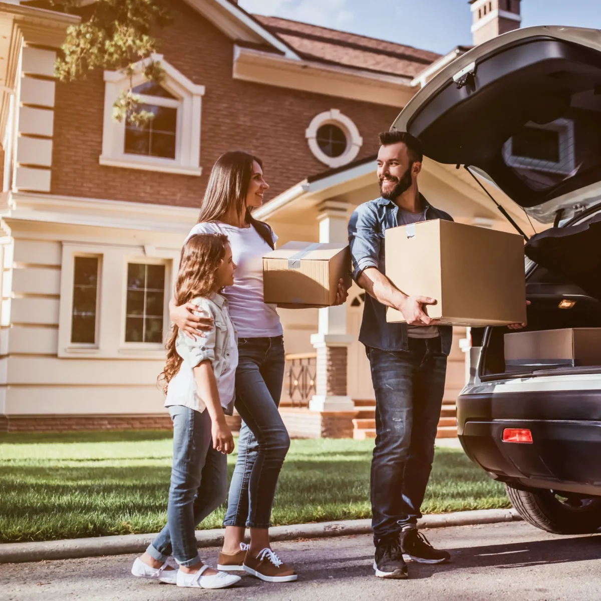 A man, woman, and young girl smile while carrying moving boxes, standing near a silver SUV and a large suburban home.