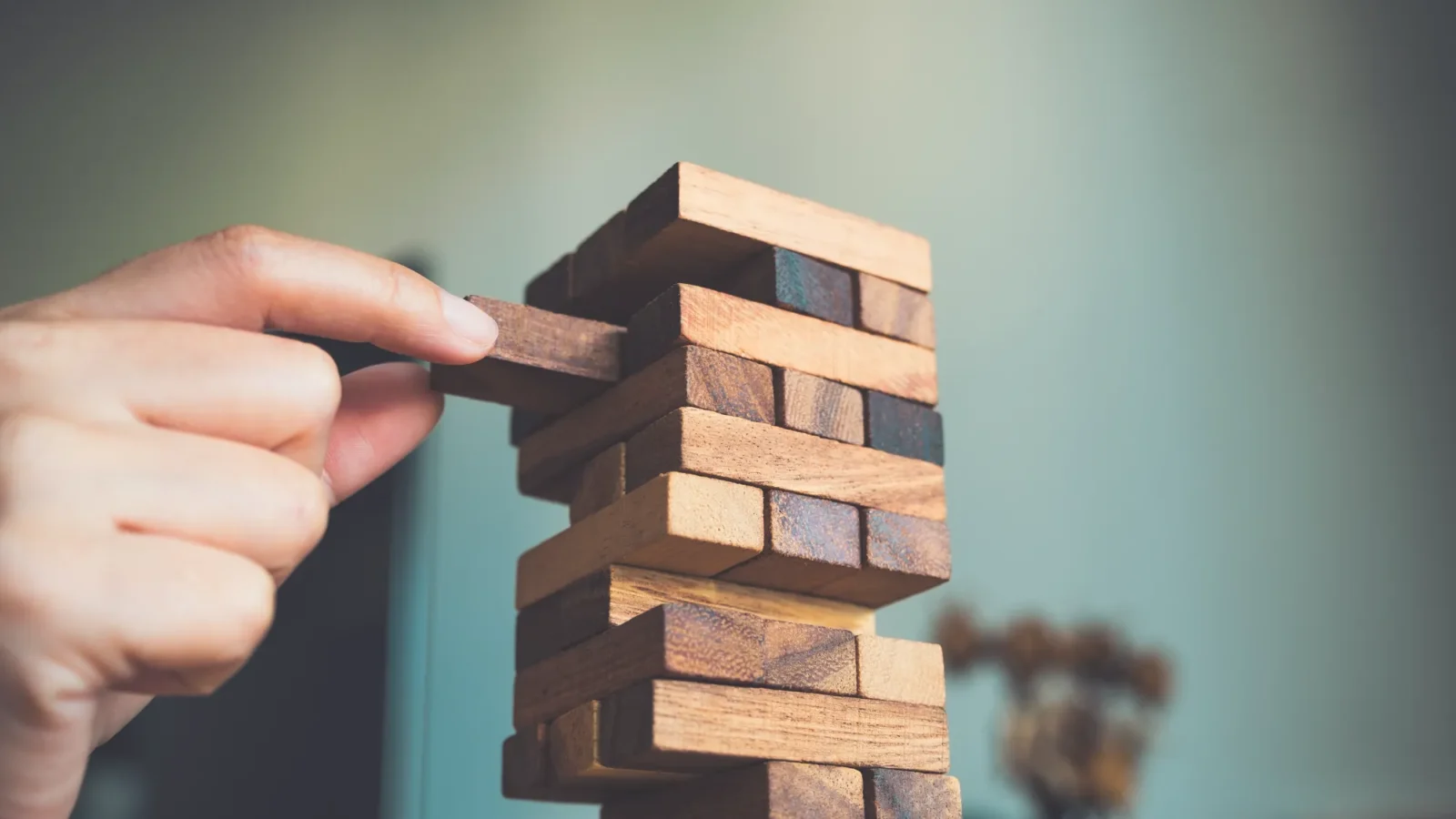 A person’s hand carefully slides a dark wooden block from the middle of a tall tower constructed of alternating wooden blocks.