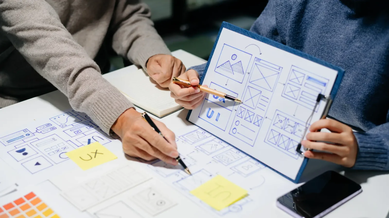 Two people collaborate on UI/UX design mockups. One points to a wireframe sketch on a clipboard, while the other draws more elements on the table surface.
