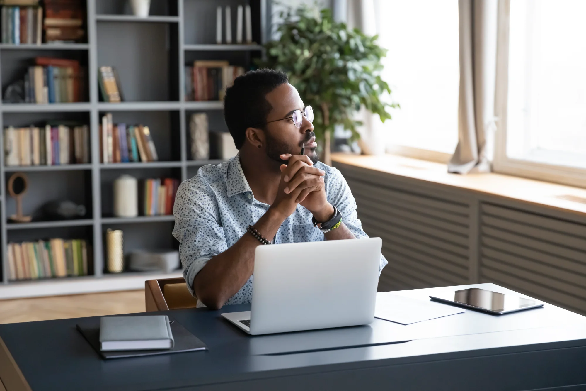A man with glasses and a patterned shirt sits at a desk with a laptop. He looks out a window with a thoughtful expression, hands clasped under his chin.