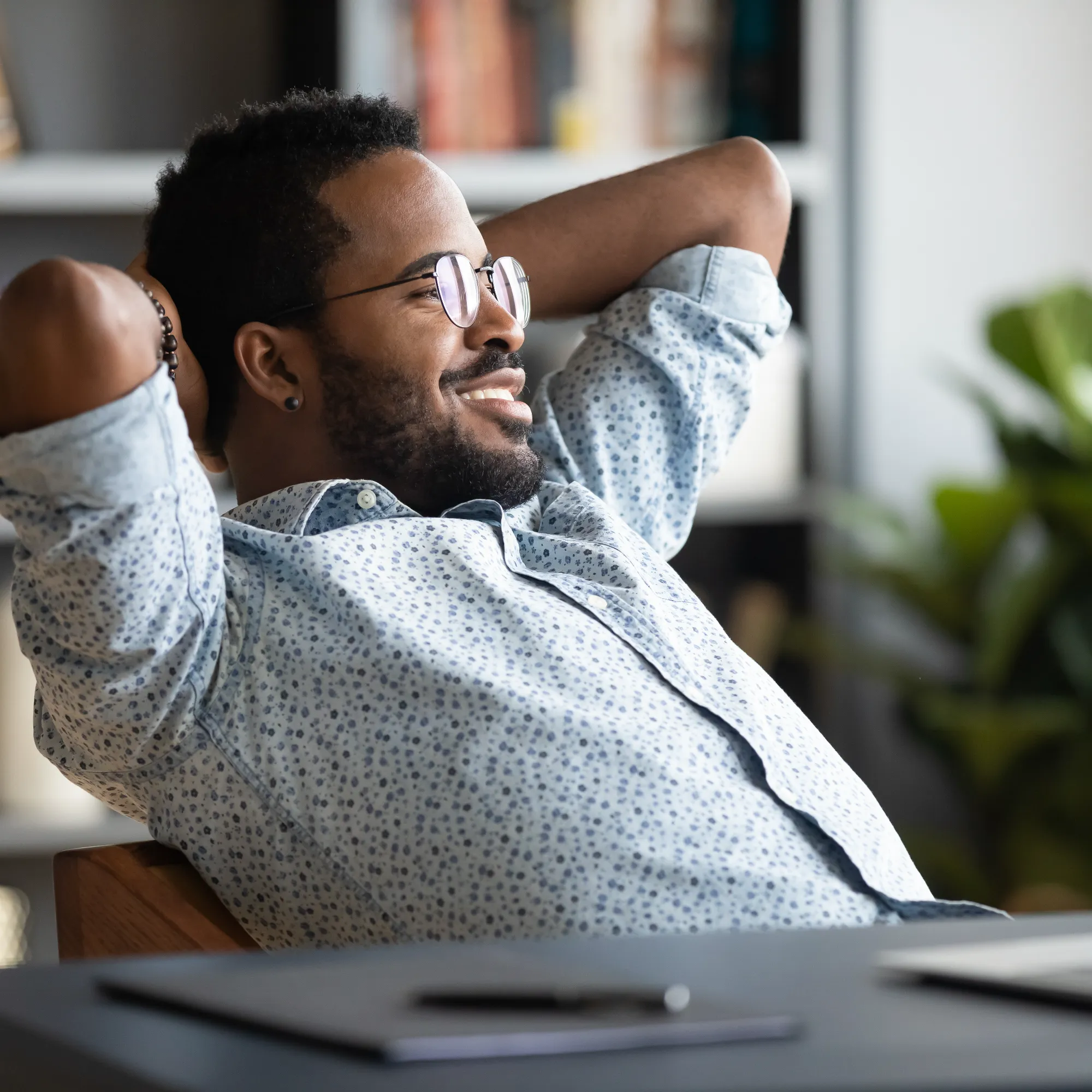 A smiling man in an office setting relaxes, leaning back in his chair with his hands behind his head, showing satisfaction.