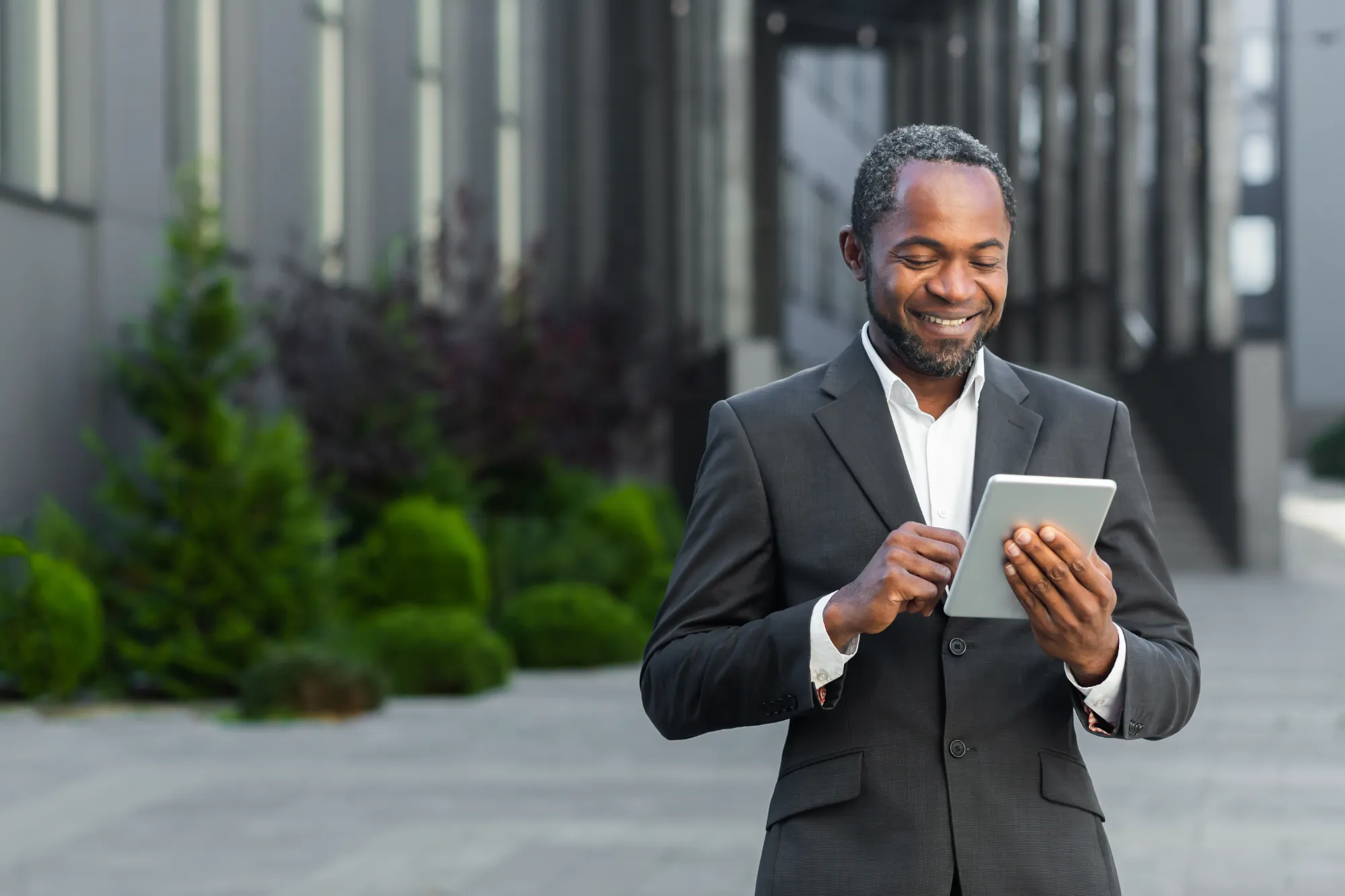A businessman in a dark suit stands outside a modern office building, smiling happily as he interacts with a silver tablet.