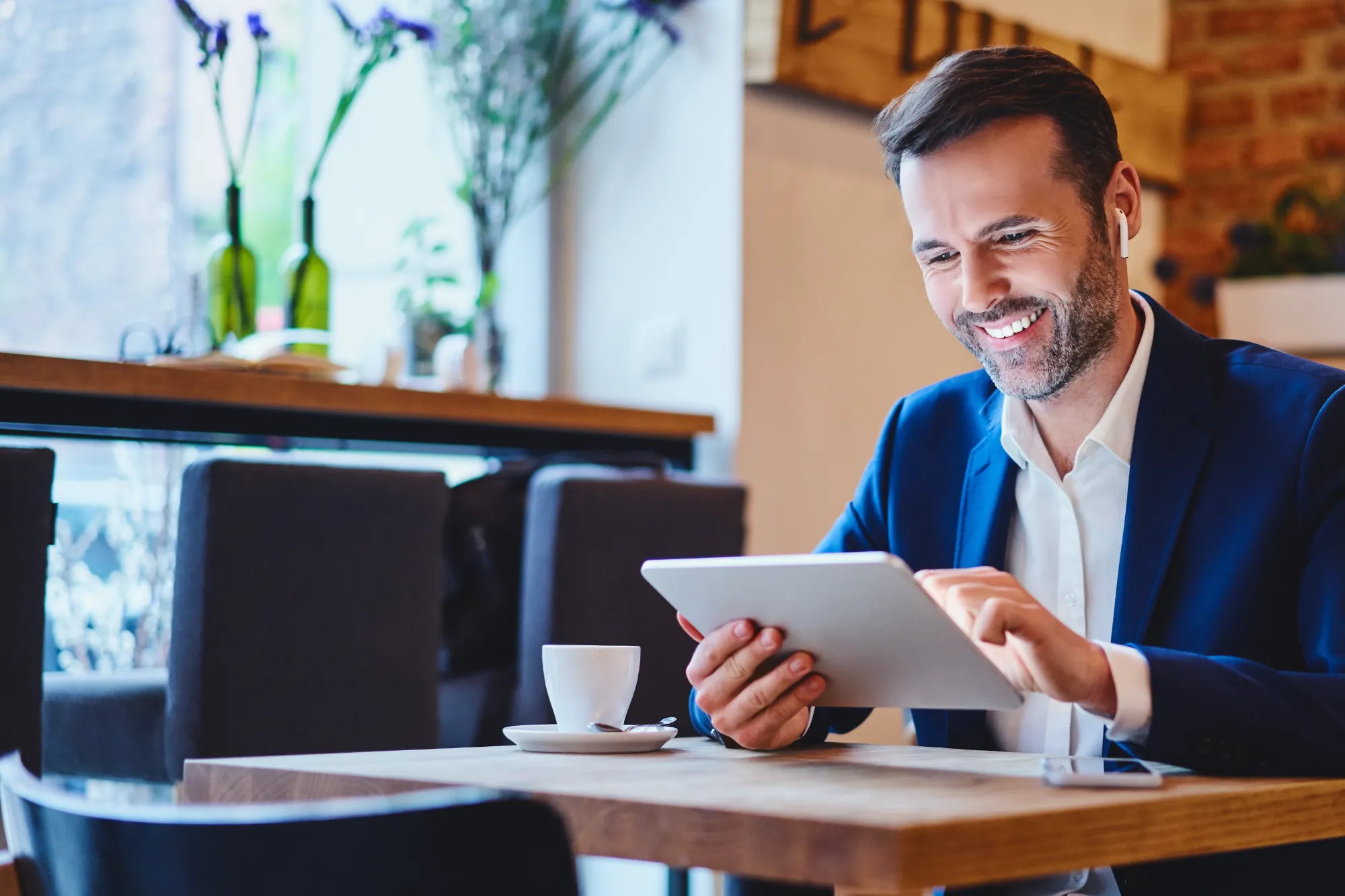 A smiling professional man in a blue suit uses a tablet and a wireless earbud while sitting at a table in a bright coffee shop.