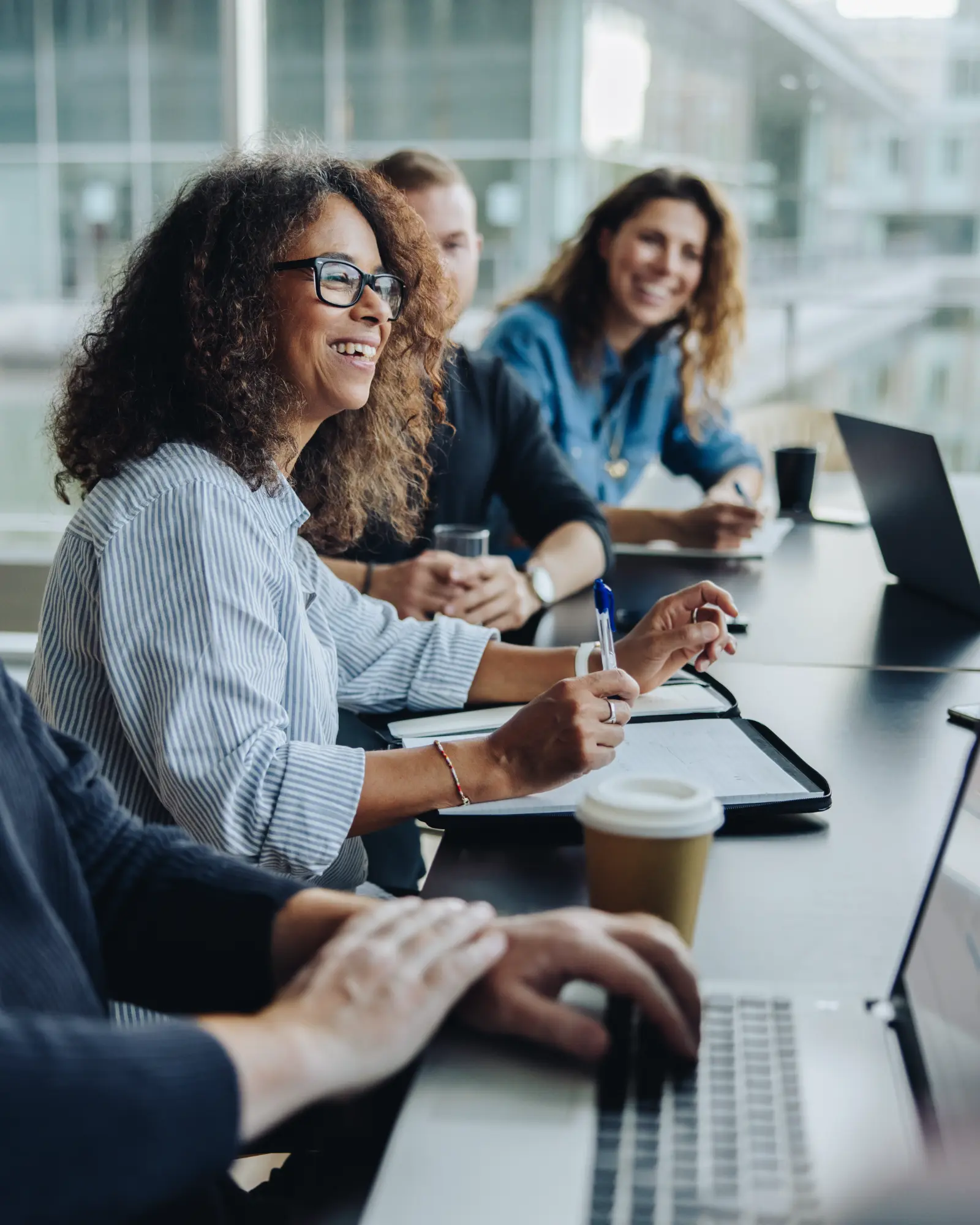 A diverse group of professionals smile happily during a meeting in a bright conference room. The main subject laughs while holding a pen and taking notes.