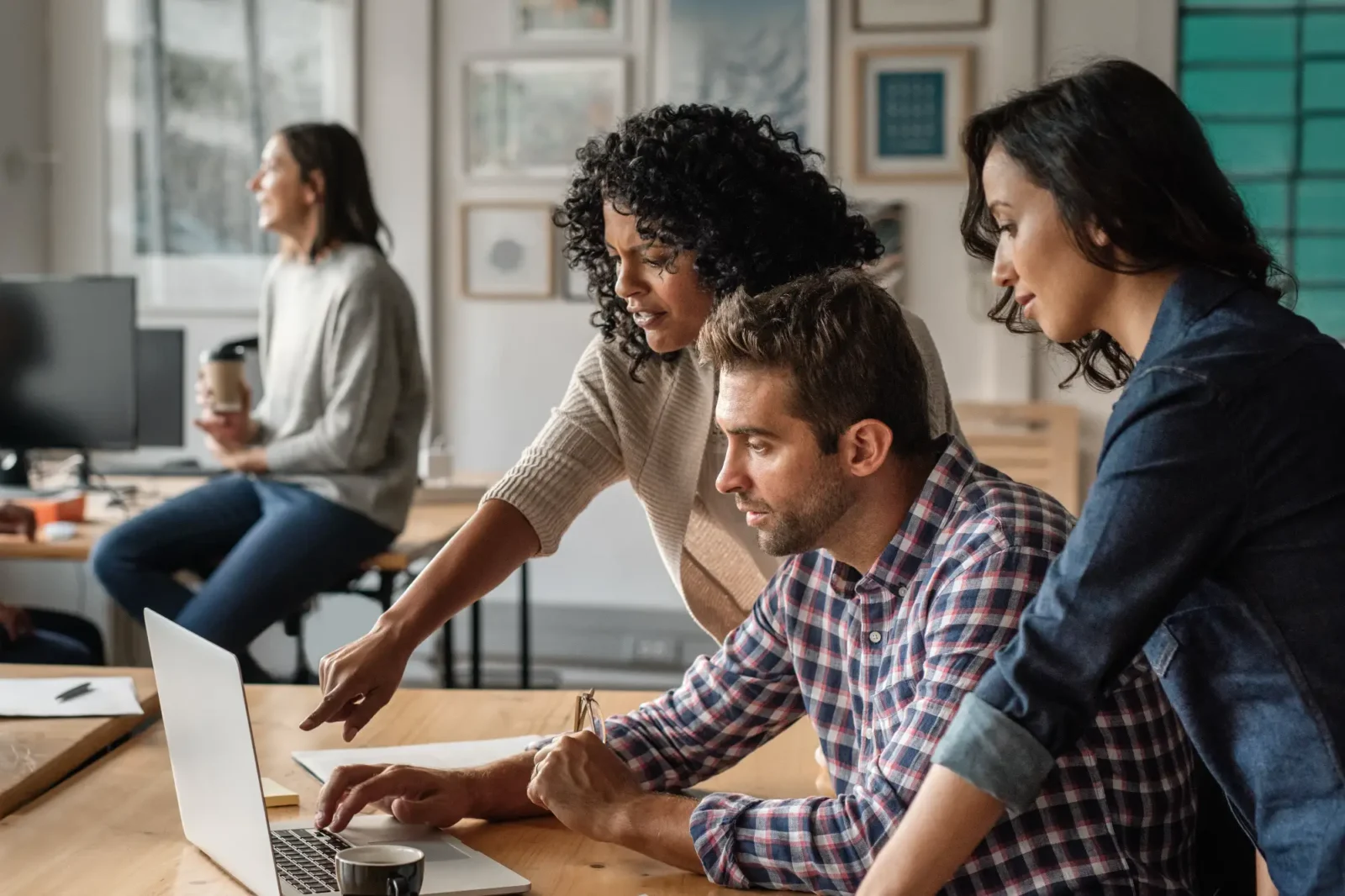 Three focused colleagues collaborate in an office, leaning over a laptop. One woman points to the screen, directing the seated man.