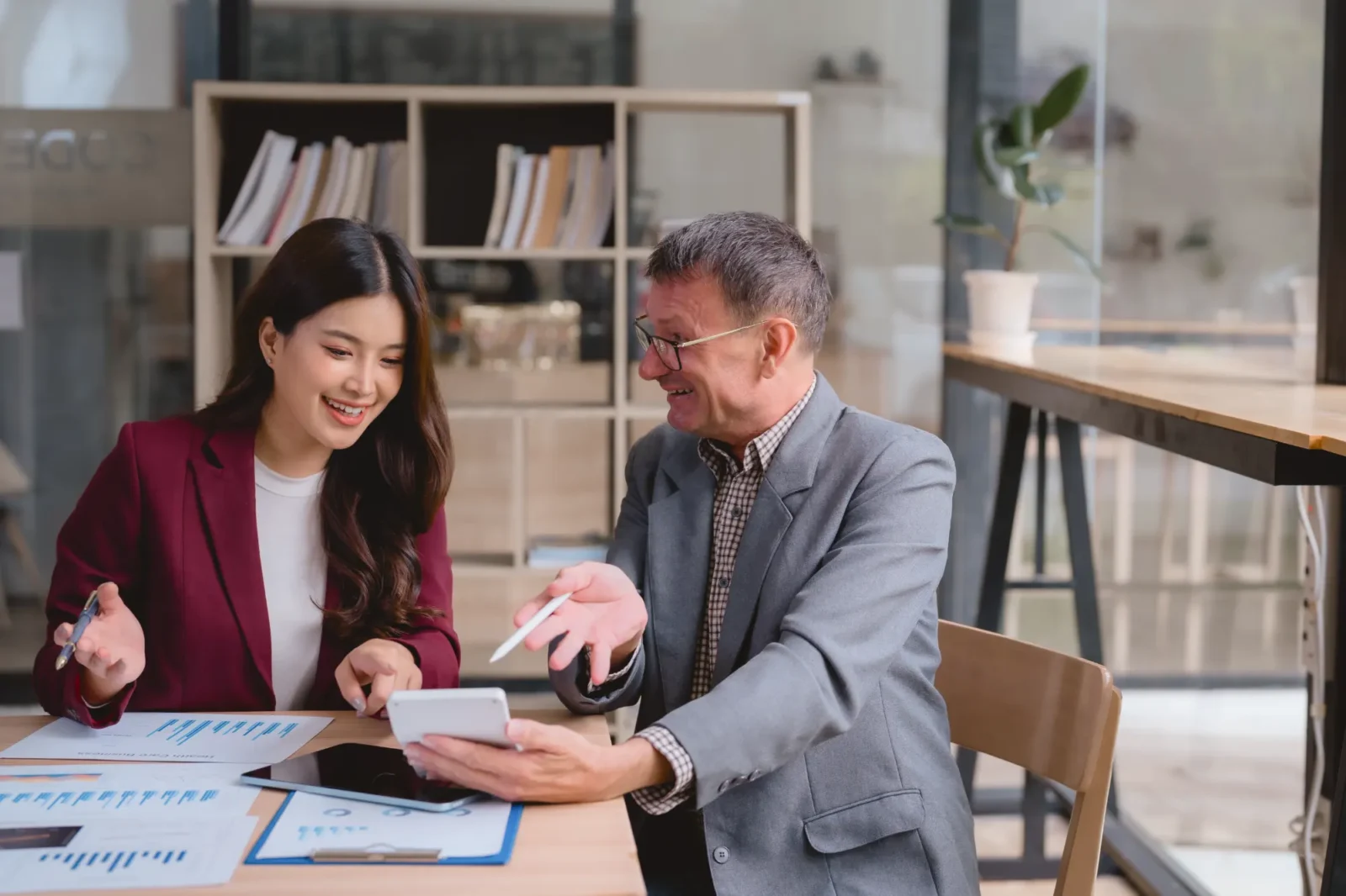A smiling man in a grey suit holds a stylus while discussing documents with a woman in a maroon blazer. They are seated at a desk covered in charts.