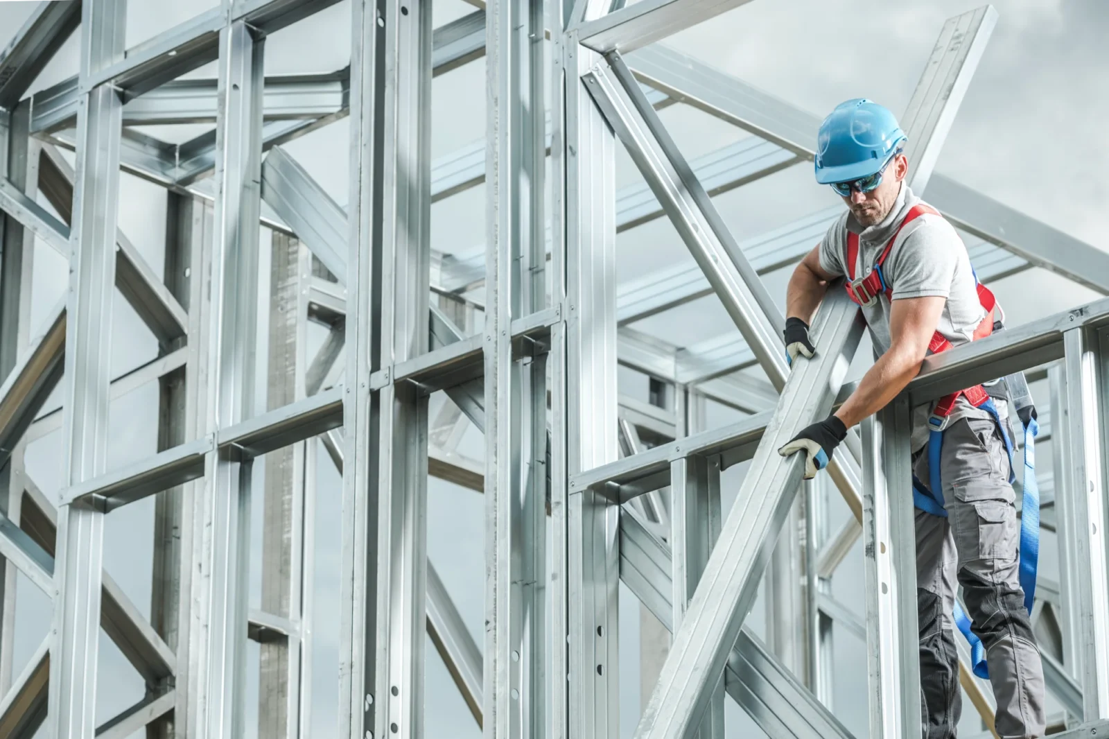 A construction worker wearing a blue hard hat and safety harness is actively installing a piece of metal framing into a large steel structure against a cloudy sky.