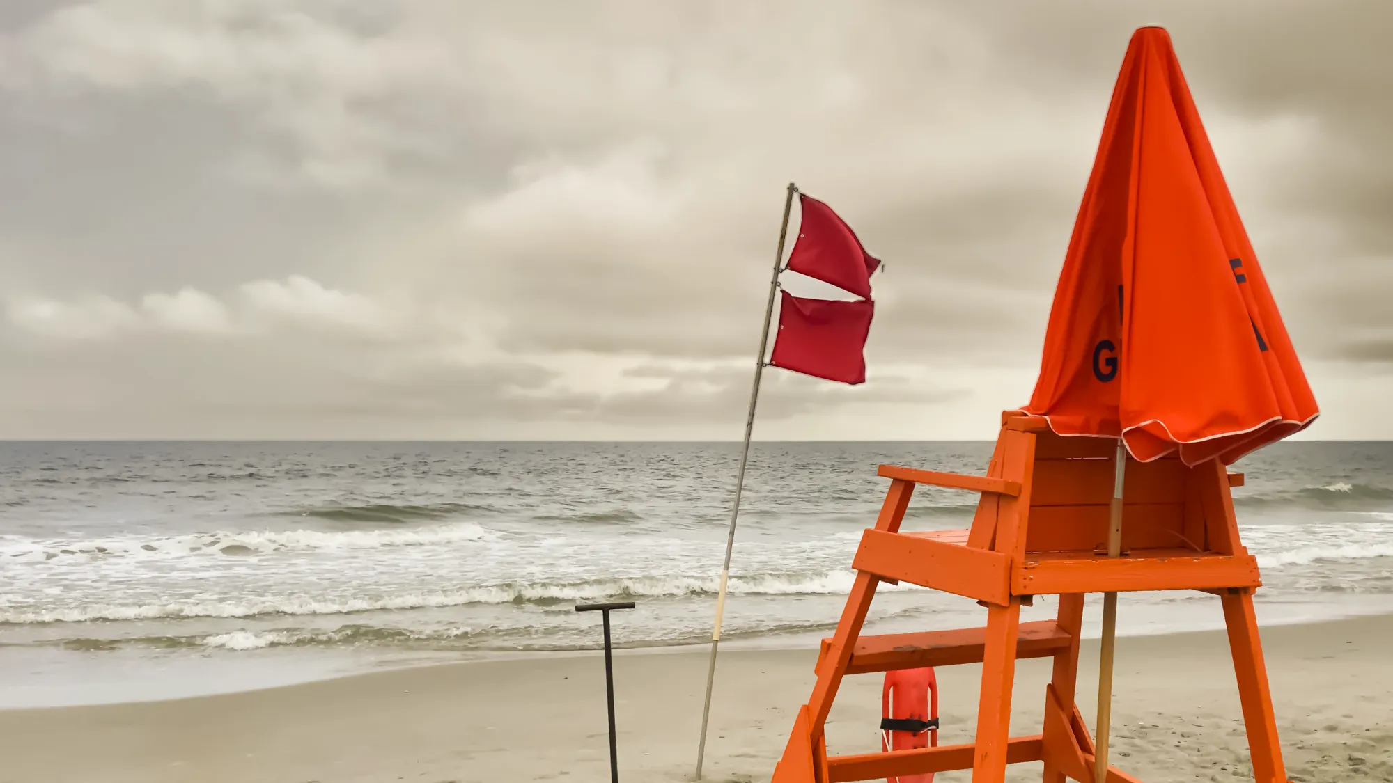 An orange lifeguard chair on a stormy beach flies two red warning flags, indicating hazardous water conditions under a cloudy sky.