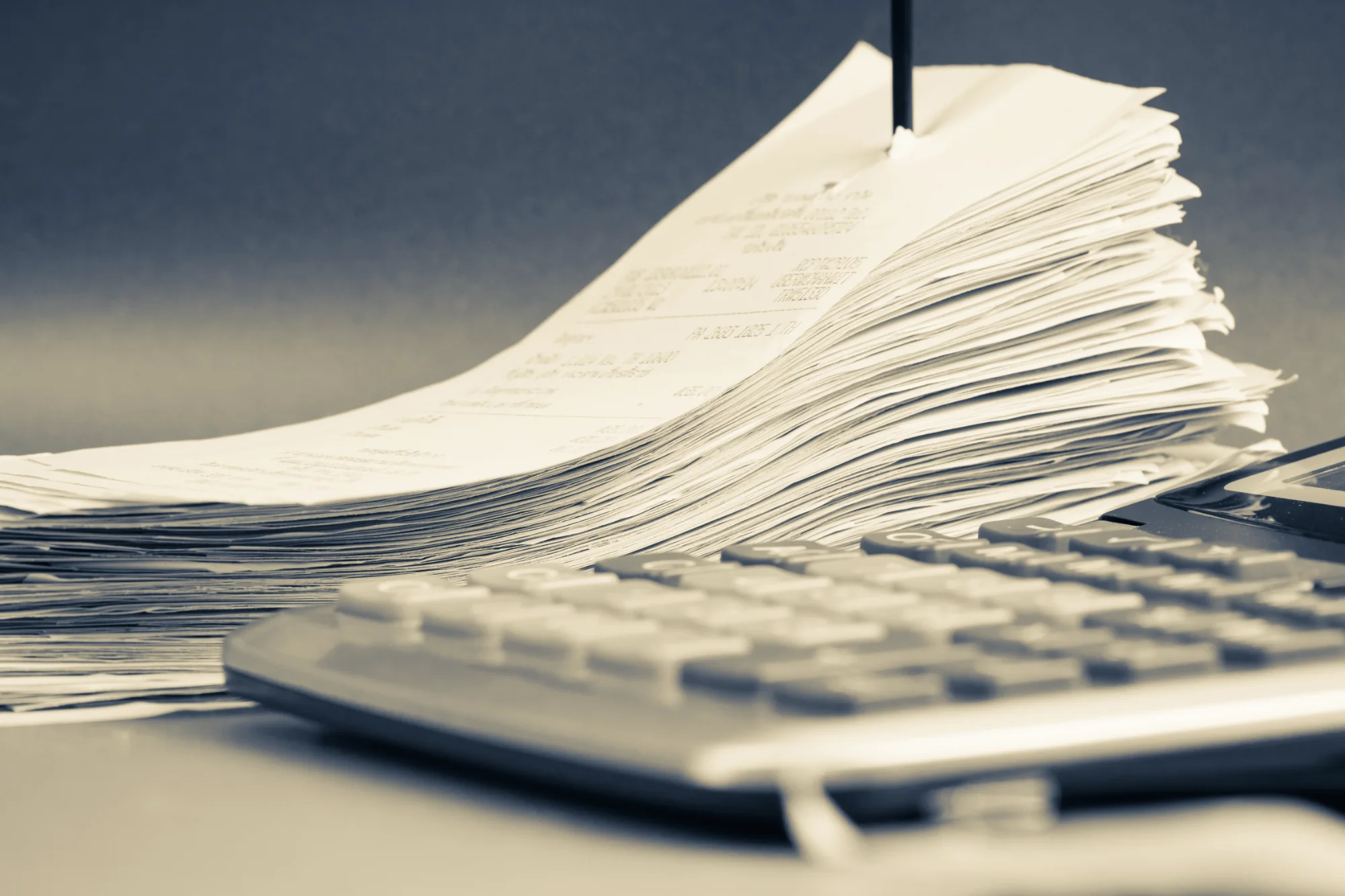 A thick stack of paper receipts, pinned by a black spike, sits next to the keyboard of a calculator in a sepia-toned image.