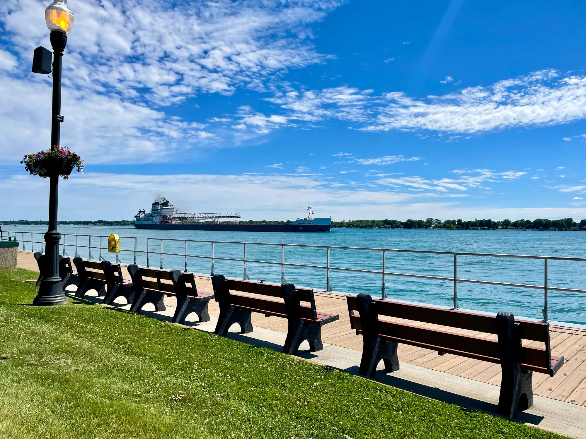 A sunny waterfront view featuring a row of dark park benches along a wooden deck next to a grassy lawn. A large freighter navigates the bright blue water under a partly cloudy sky.
