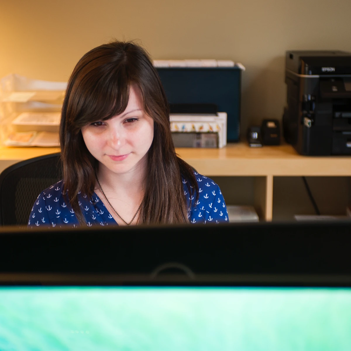 Sara, a young woman with long brown hair and a focused expression, sits at her office desk working on a computer.