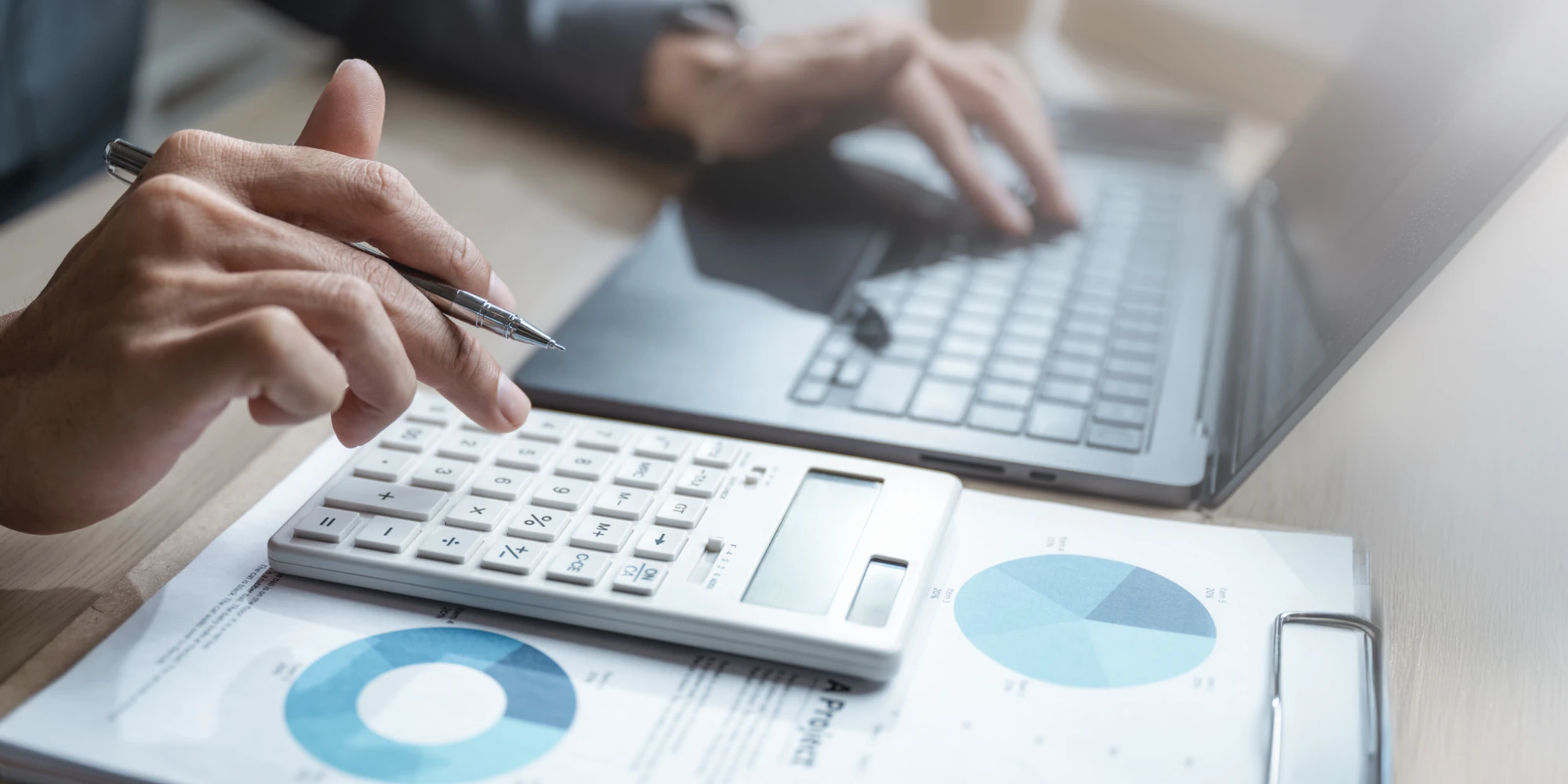 A hand holds a pen above a calculator resting on documents displaying blue pie charts, while a second hand types on a laptop.