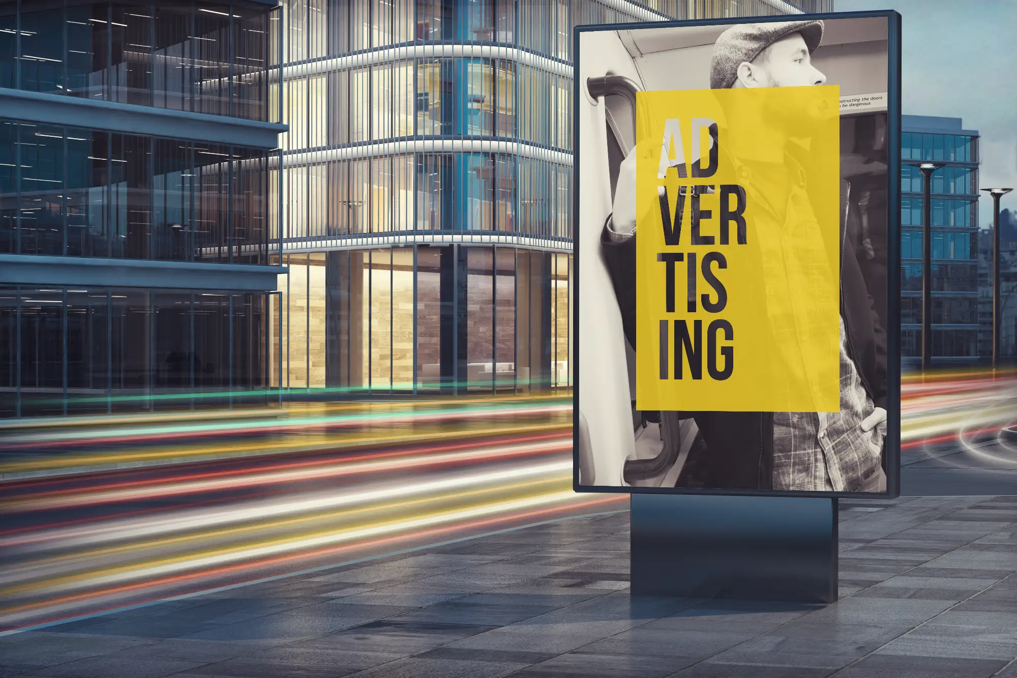 A vertical digital sign on a city sidewalk displaying the word "ADVERTISING" over a man in a flat cap. Streaks of light from passing traffic blur across the modern glass buildings in the background.